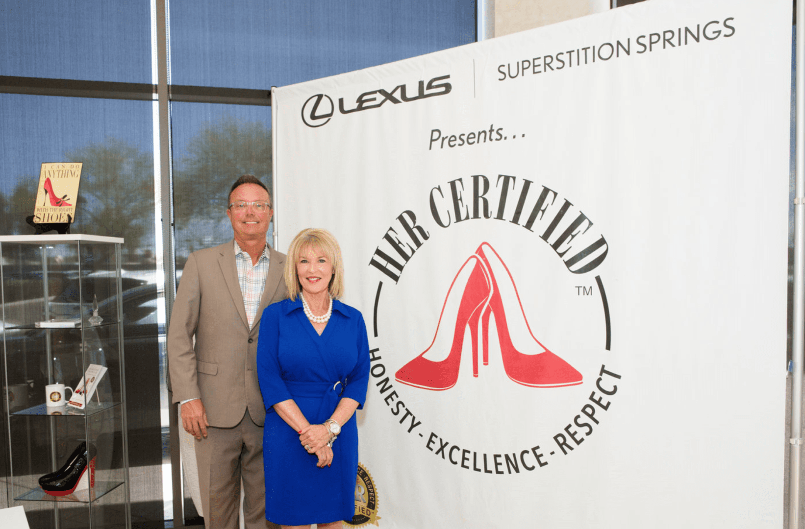 Two individuals, a man and a woman, standing in front of a promotional banner that reads "lexus superstition springs presents: mr. certified" with a red high heels logo.