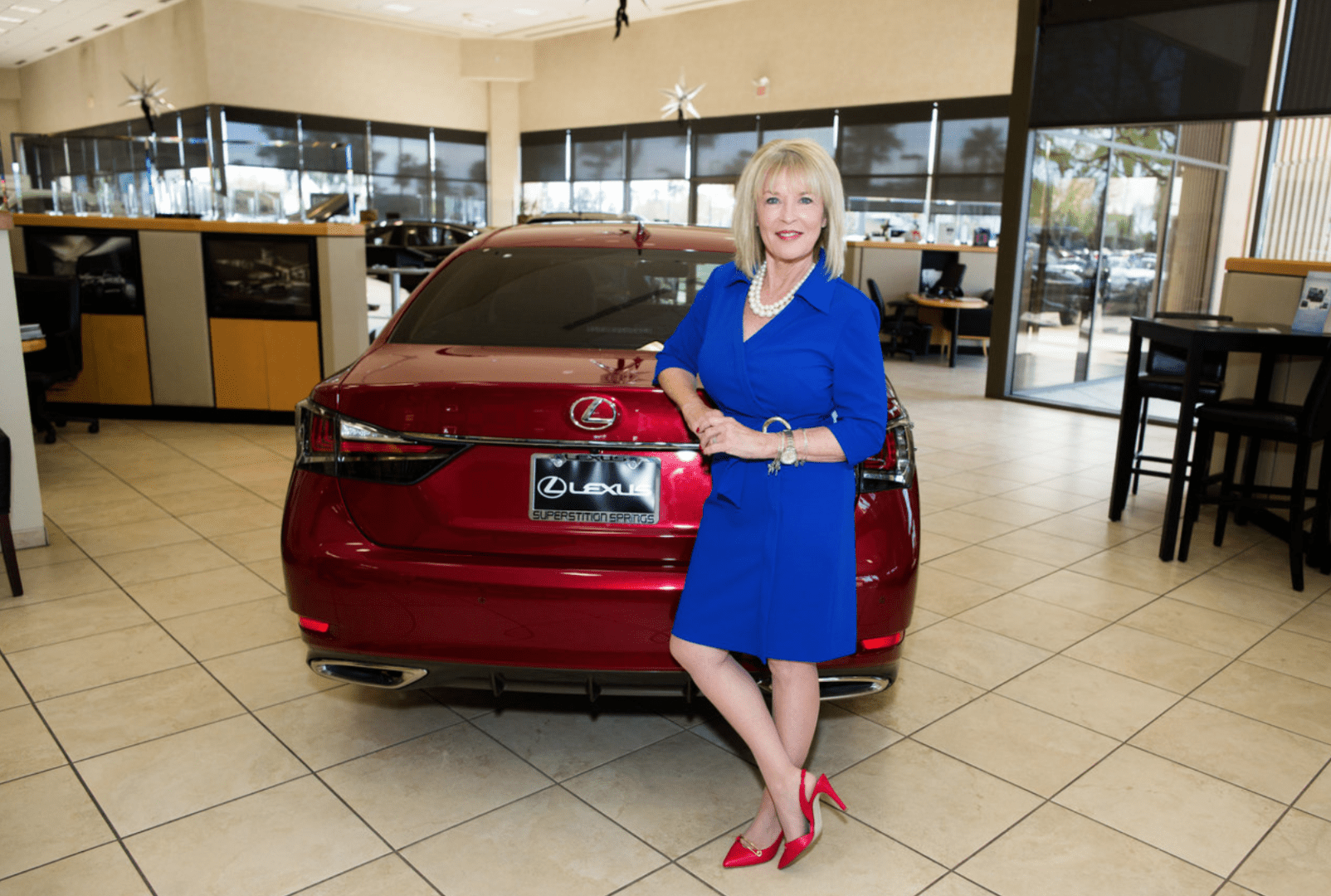 A woman in a blue dress and red heels stands confidently in front of a red lexus in a car dealership showroom.
