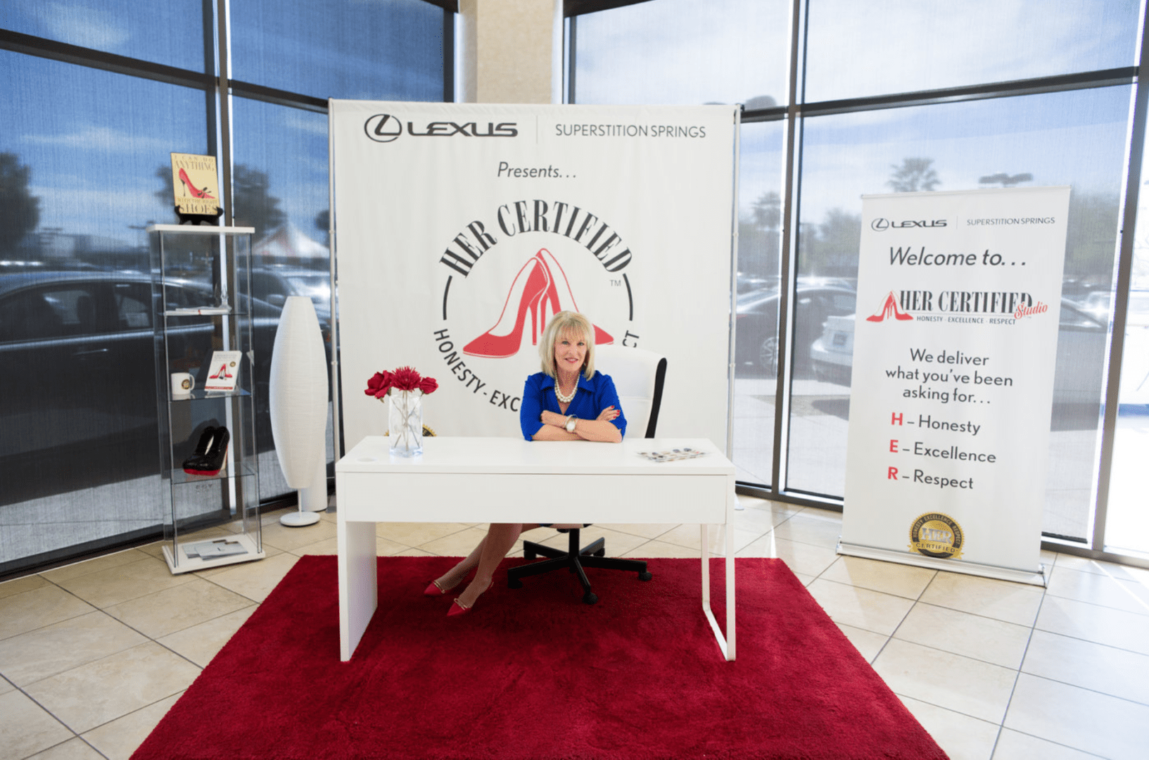 A woman sits at a promotional table with a lexus superstition springs banner in a dealership, smiling at the camera.