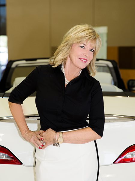 A woman with blonde hair in a black shirt smiles, leaning on the trunk of a white convertible car in a showroom.