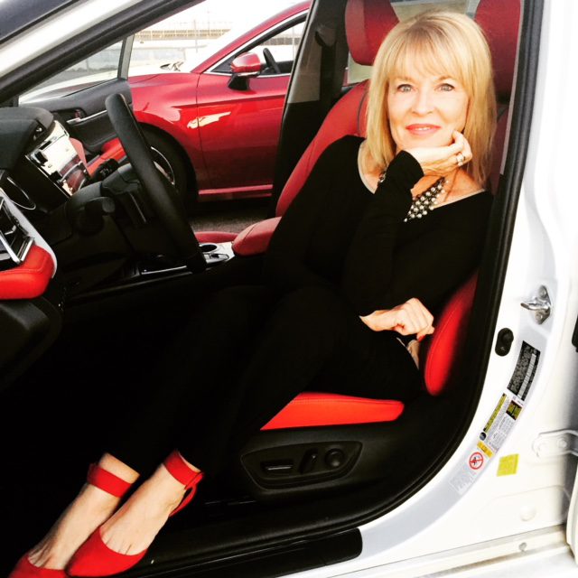 A mature woman with blonde hair, wearing a black outfit and red shoes, sitting inside a car with red and black interior, smiling at the camera.