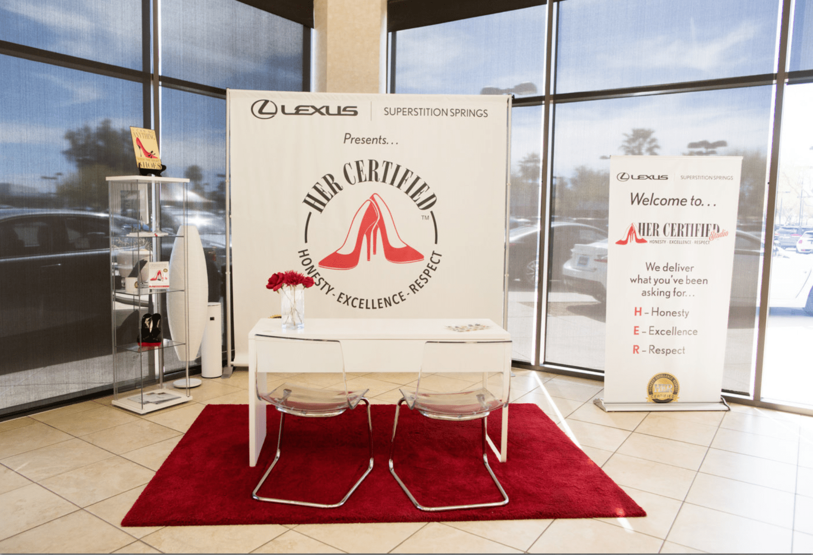 A promotional booth for the "her certified" program by lexus, featuring a banner and information stand, with two chairs on a red carpet in a dealership.