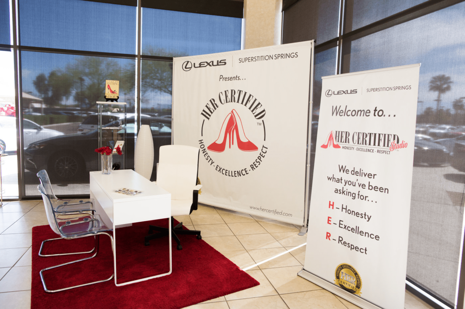 A dealership lobby with a lexus banner, two promotional stands, and a seating area with a white table and chairs on a red carpet.
