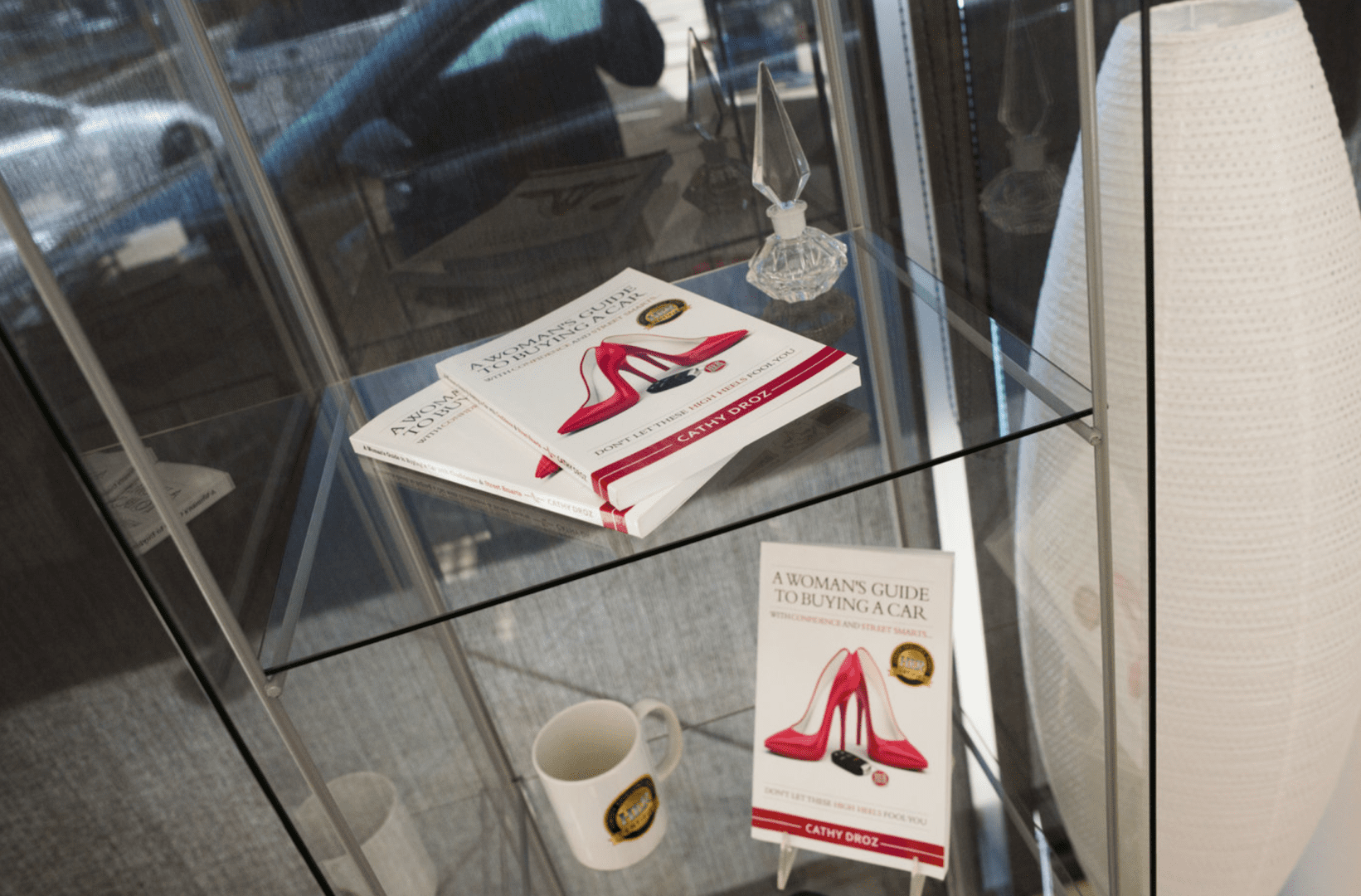 A book titled "a woman's guide to buying a car" by cathy droz is displayed on a glass shelf near a white mug.