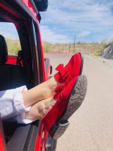 A person reclines in a car, sticking their legs out the window, showcasing bright red shoes with bows against a backdrop of a rocky landscape.