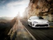 A white sports car speeds along a winding mountain road at sunset, with a sharp cliff on one side and panoramic views in the background.