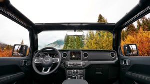 Interior view from a jeep’s driver seat showing the steering wheel and dashboard with a scenic autumn forest and mountain landscape visible through the windshield.