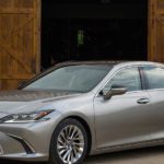 A silver lexus sedan parked in front of a large wooden barn with doors open, surrounded by greenery.