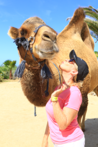 A woman standing next to a camel on the beach.