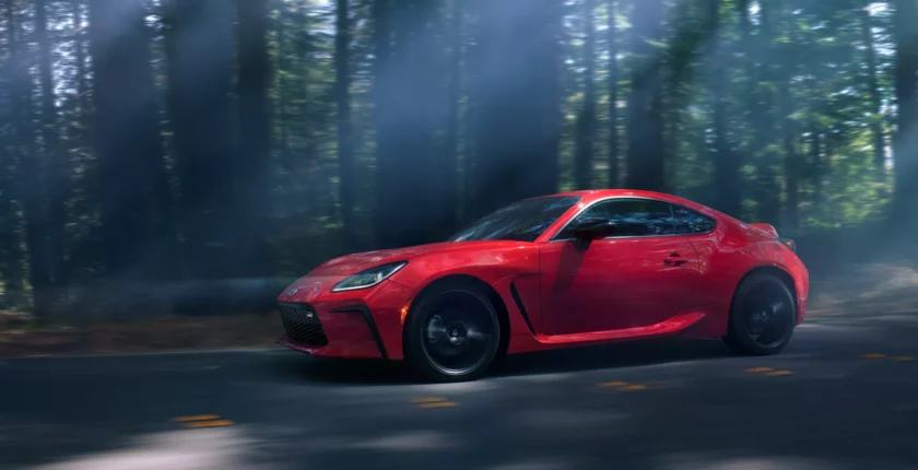 A red sports car driving down the road in front of trees.