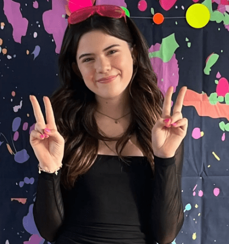 Young woman in a black outfit and pink headband making a peace sign with both hands, smiling in front of a colorful splattered paint backdrop.